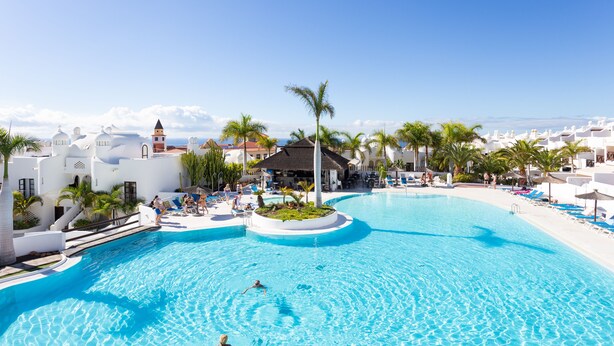 A large swimming pool with clear blue water, surrounded by white buildings and palm trees.