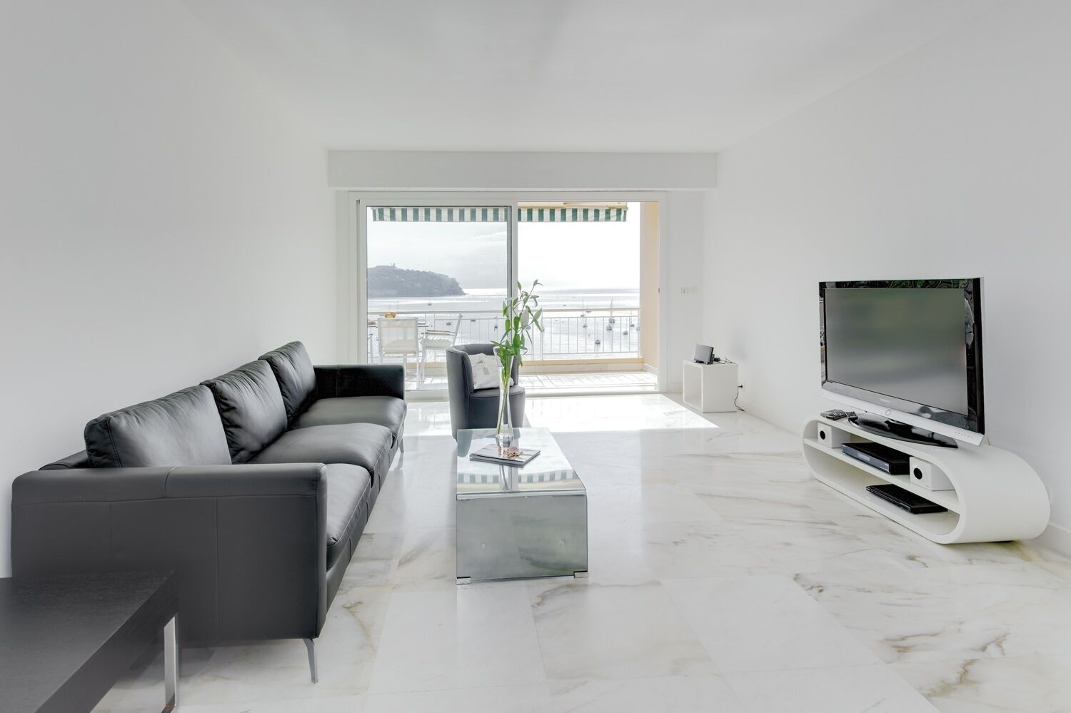 A modern living room with a black leather sofa, a glass coffee table, a white entertainment unit with a flat-screen TV, and a view of the sea through large windows.
