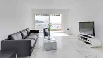 A modern living room with a black leather sofa, a glass coffee table, a white entertainment unit with a flat-screen TV, and a view of the sea through large windows.