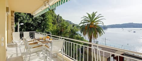 A balcony with white chairs and a table, overlooking a body of water with boats and greenery.
