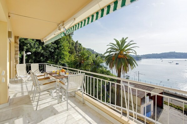Un balcon avec des chaises blanches et une table, donnant sur une étendue d’eau où l’on peut voir des bateaux et de la végétation.