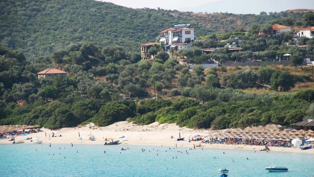A coastal area with a beach, boats, and buildings on a hillside. - Demi Studios (Aristotelis)