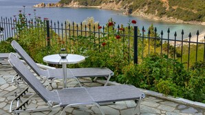 A terrace with two lounge chairs and a table, overlooking a coastal landscape with a fence and blooming flowers. - Demi Studios (Aristotelis)