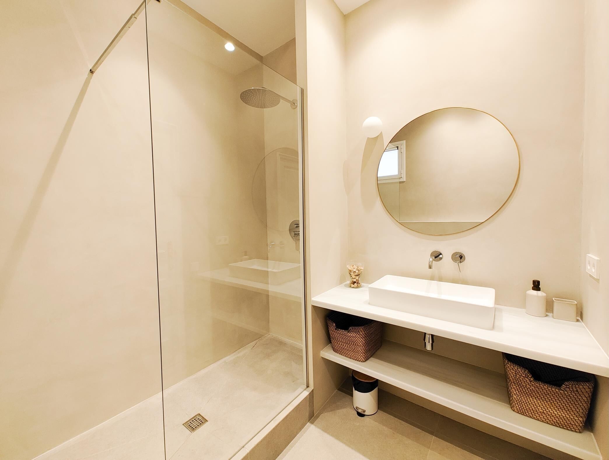 A modern bathroom with a glass-enclosed shower, a round mirror, and a white sink with storage shelves.