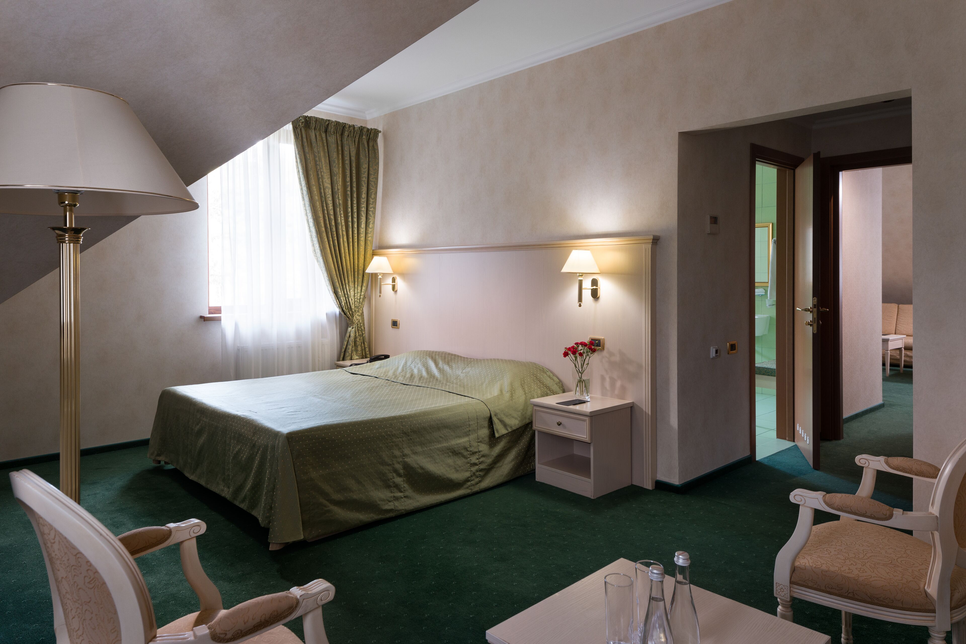 A hotel room with a bed, bedside lamps, a nightstand, a chair, a table, and a vase with flowers.
