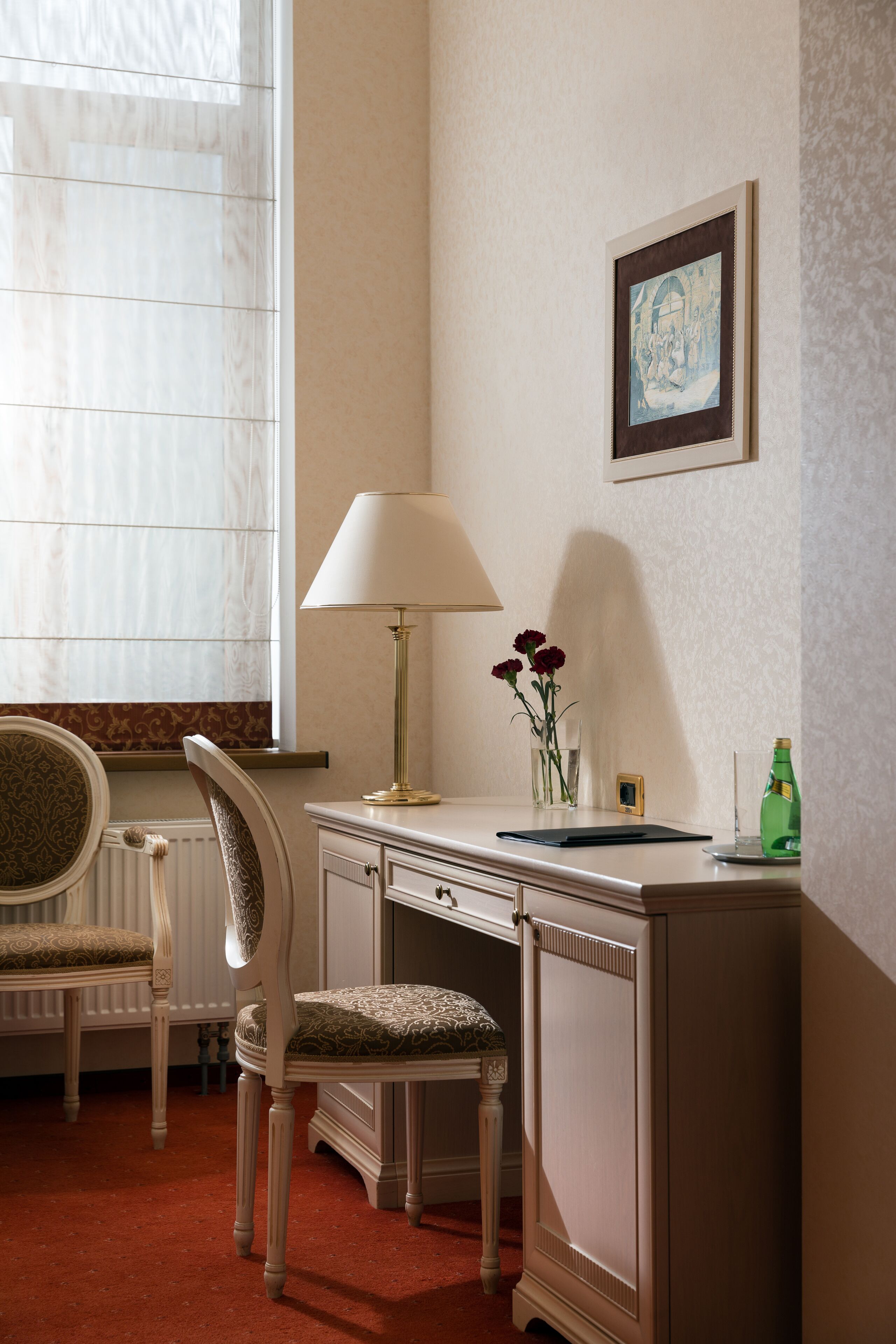 Ett rum med ett skrivbord, en stol, en lampa, en vas med blommor och en inramad tavla på väggen.