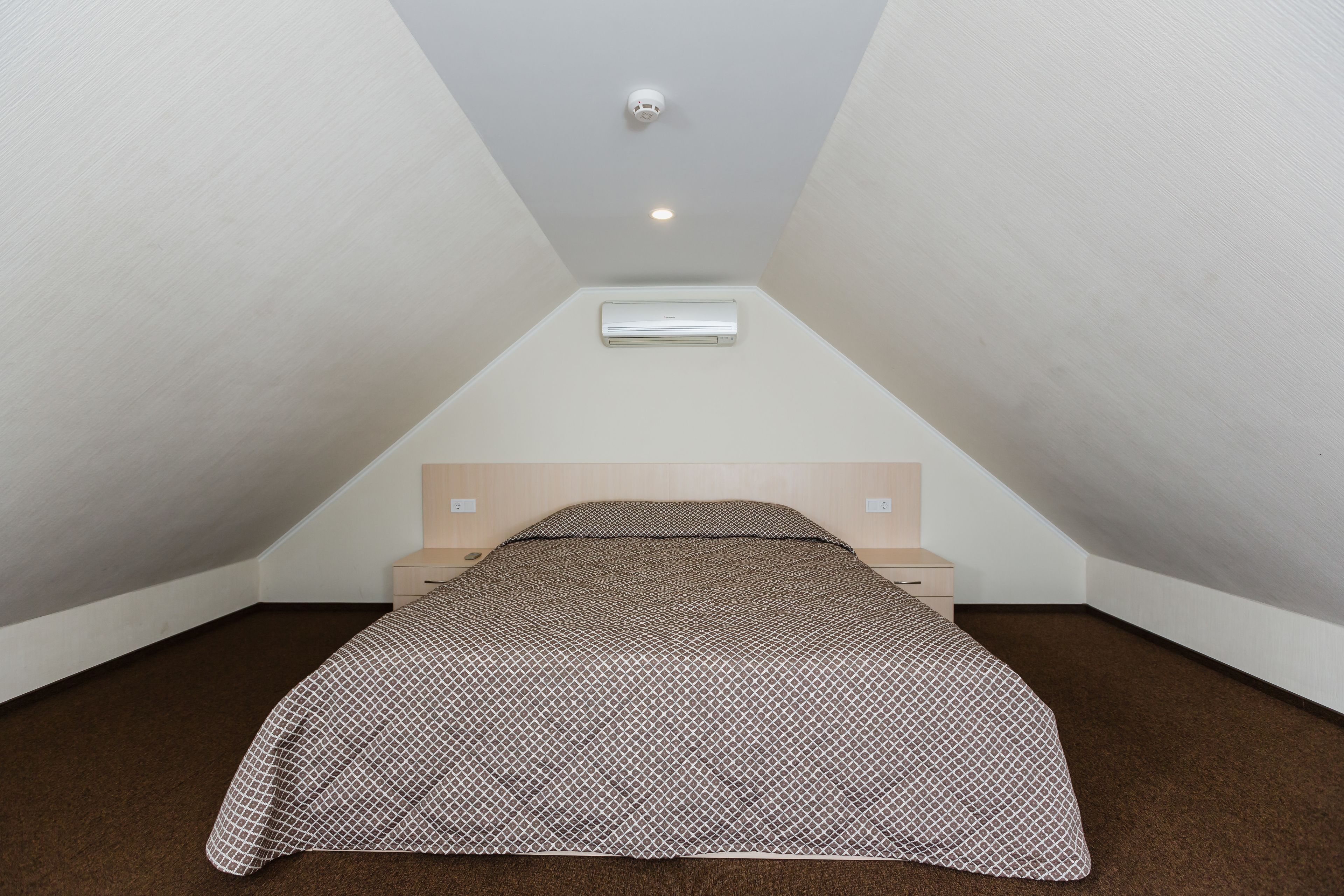 A small, triangular attic bedroom with a bed, a nightstand, and an air conditioning unit on the wall.