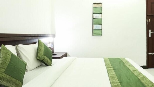 A hotel room with a bed, pillows, a nightstand, a wall-mounted light switch, and a door.