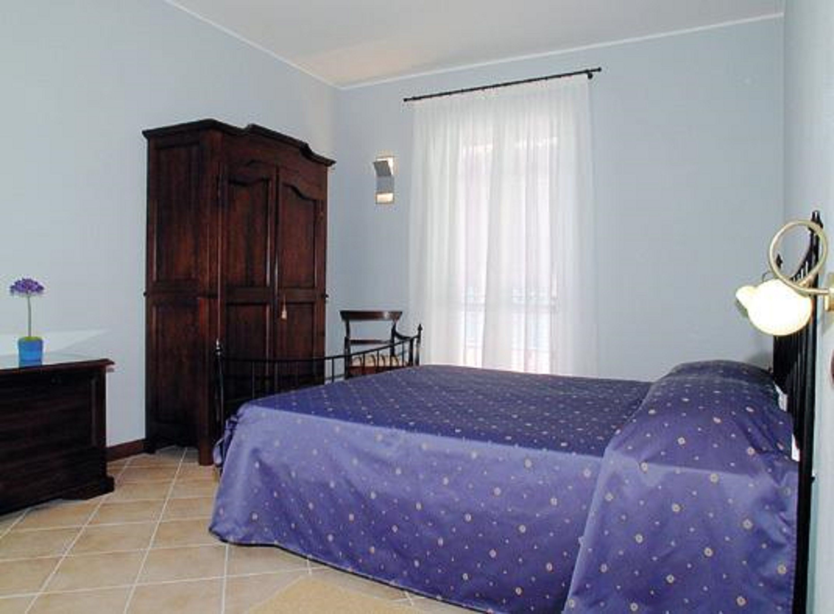 Une chambre à coucher comprenant un lit, une armoire en bois, une chaise, une petite table avec un vase et une fenêtre avec des rideaux.