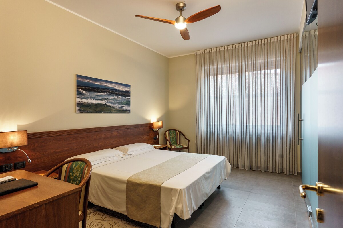 A hotel room with a bed, a desk, a chair, a ceiling fan, a painting on the wall, and a window with curtains.