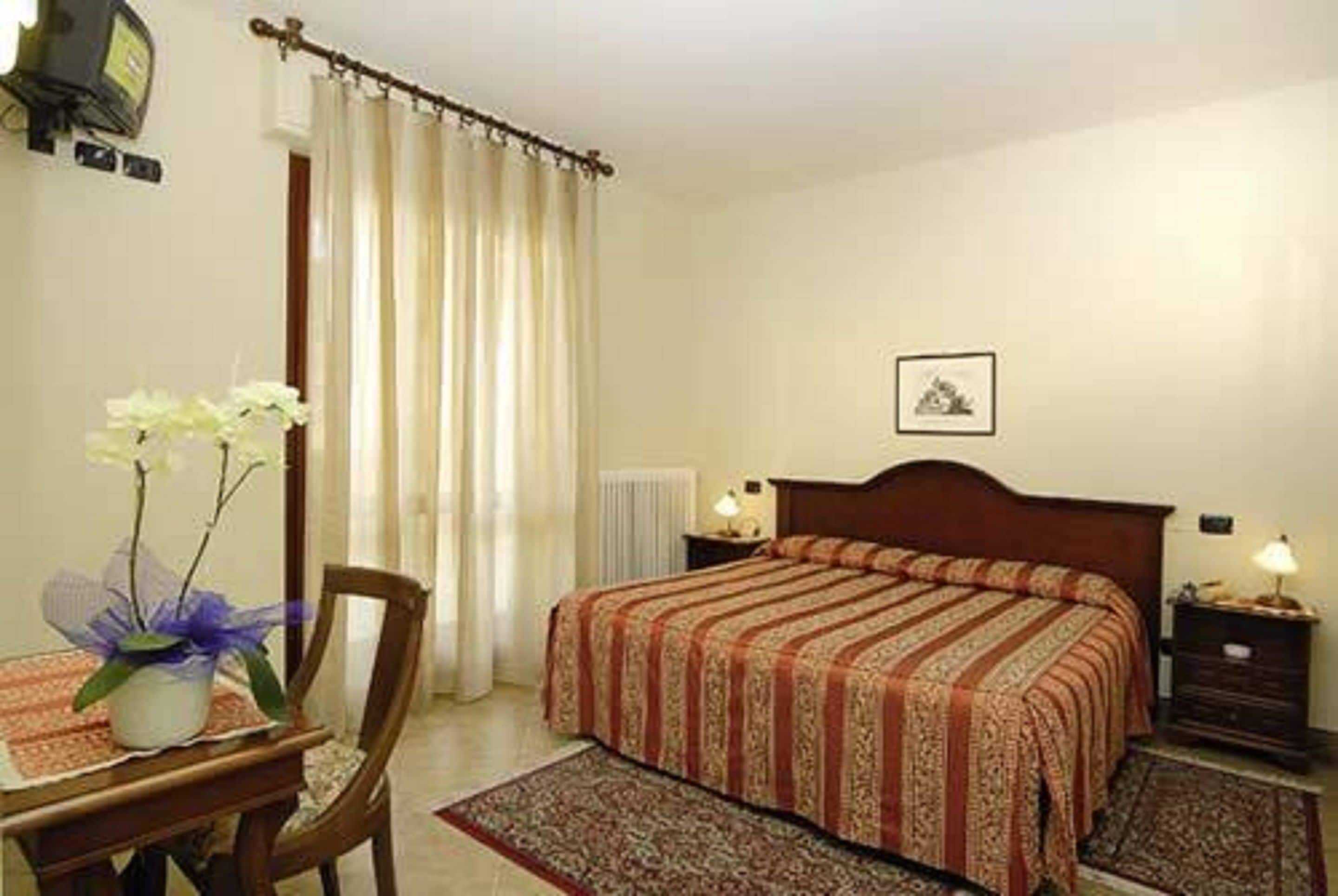 A hotel room with a bed, a chair, a table with a vase of flowers, a nightstand, and a picture on the wall.