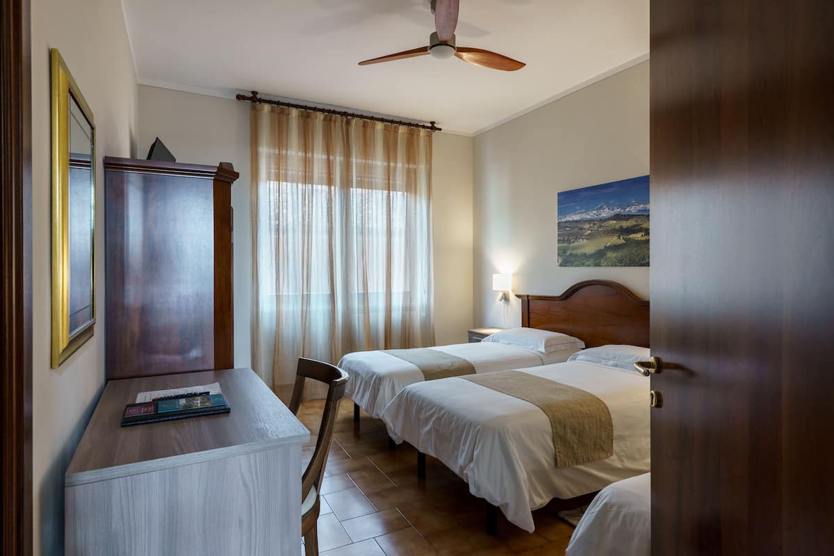 Ein Hotelzimmer mit zwei Betten, einem Schreibtisch, einem Stuhl, einem Kleiderschrank, einem Deckenventilator und einem Bild an der Wand.