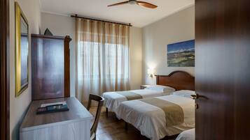 A hotel room with two beds, a desk, a chair, a wardrobe, a ceiling fan, and a painting on the wall.