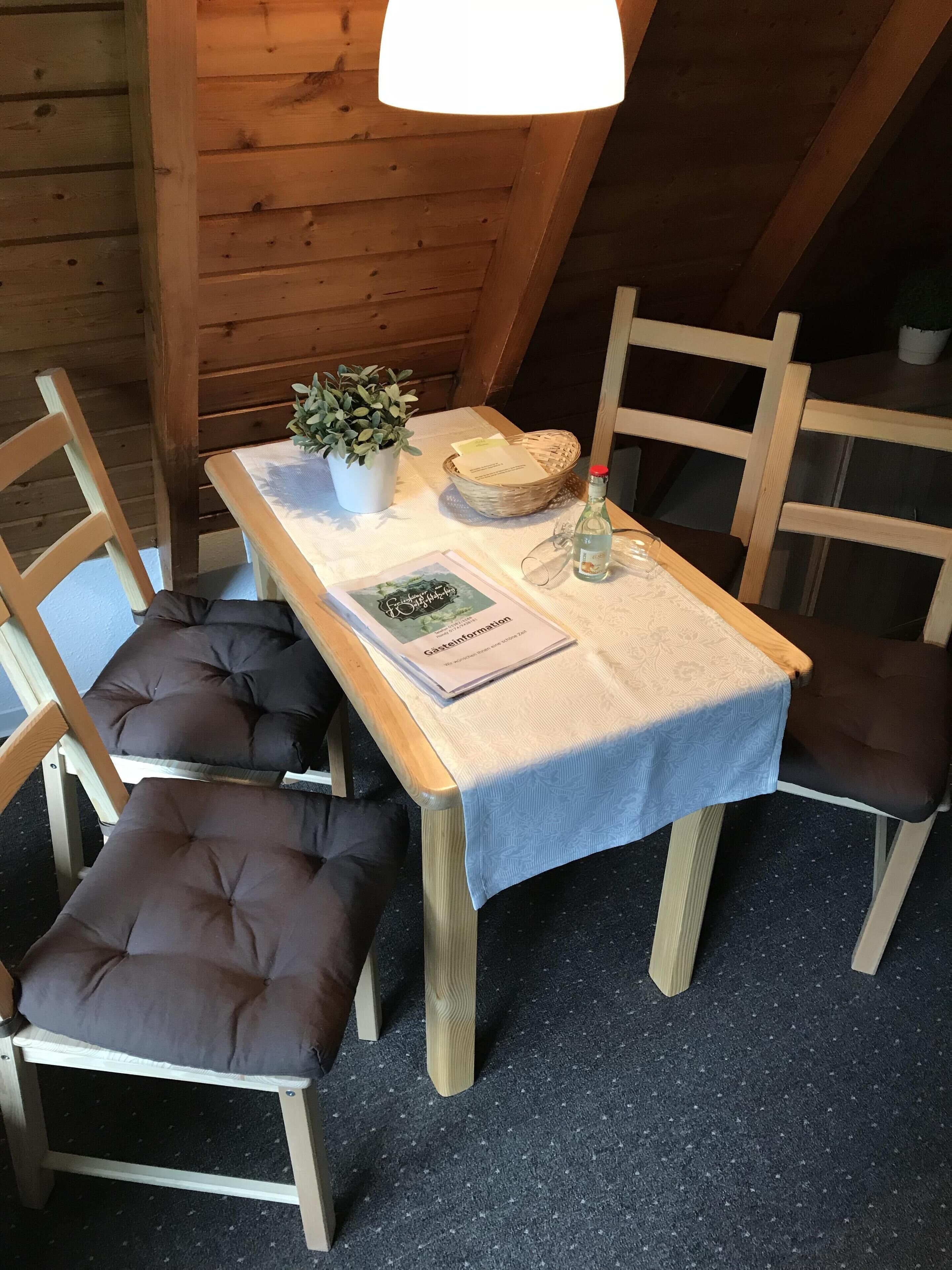 A wooden dining table with a white tablecloth, two cushioned chairs, a potted plant, a basket, a bottle, and a menu.