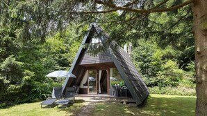 Een modern, driehoekig houten vakantiehuisje met glazen deuropening, omringd door weelderige begroeiing.
