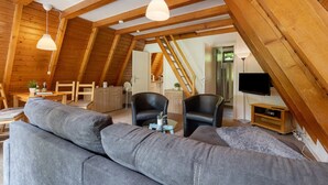 Een knus loft-woongedeelte met een hoge plafond, zichtbare houten balken, een grijze modulaire bank, twee zwarte leren fauteuils, een glazen salontafel en een flatscreen televisie op een houten dressoir.