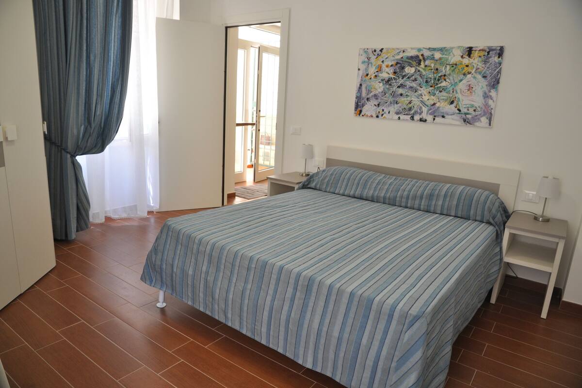 Une chambre à coucher avec un grand lit, une table de chevet, un tableau accroché au mur et une vue sur l’extérieur, visible à travers une porte ouverte.