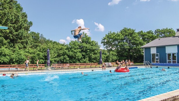 A swimming pool with people in the water and on the edge, a lifeguard tower, and a building with a blue roof in the background. - Beautiful studio in Løgumkloster (Løgumkloster)