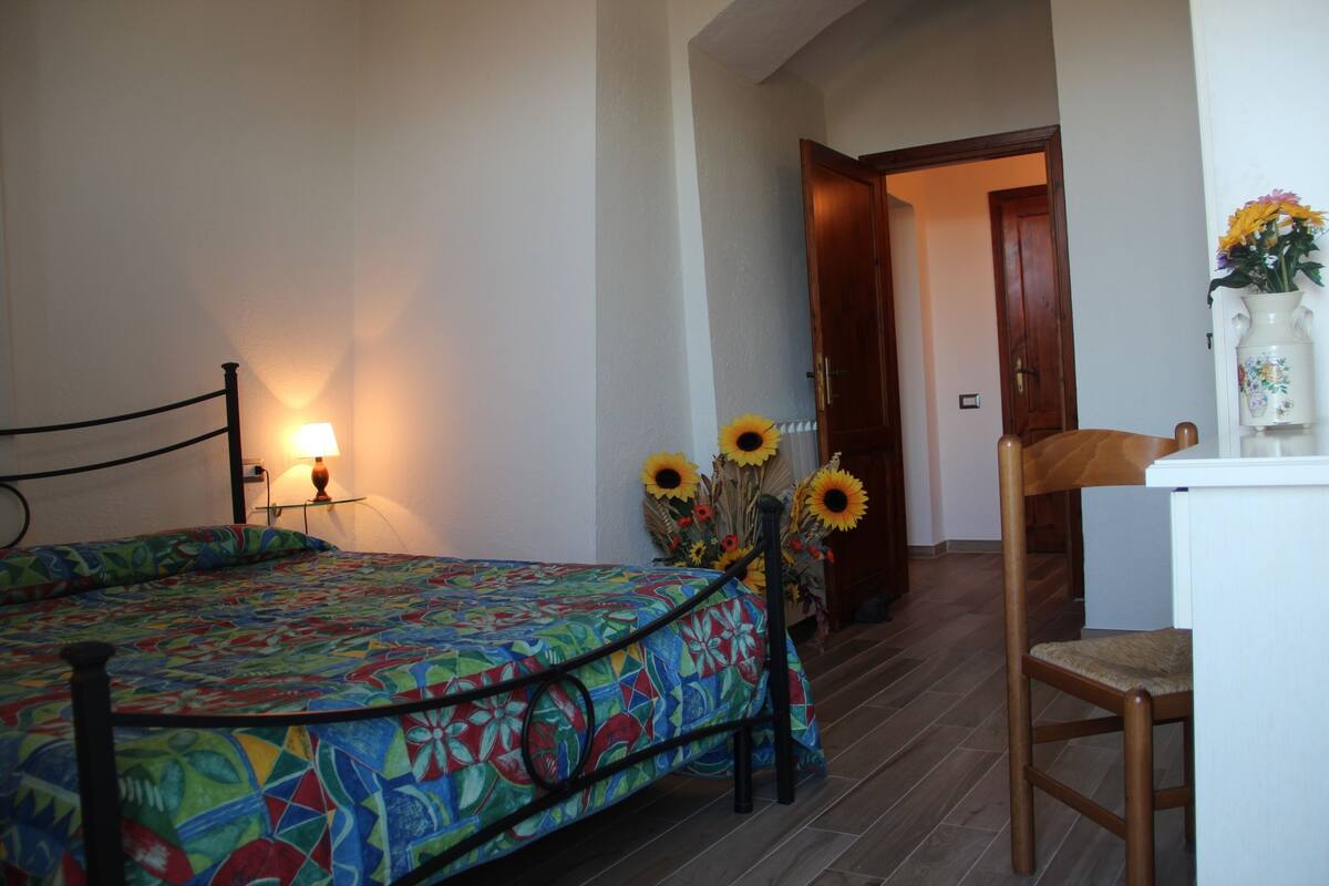 Ein Schlafzimmer mit einem Bett, einem Sessel, einem Schreibtisch und einer Vase mit Blumen.