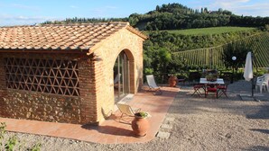 Un bâtiment rustique en pierre, doté d’un toit en tuiles, d’une terrasse avec des sièges extérieurs et d’un vignoble en arrière-plan.