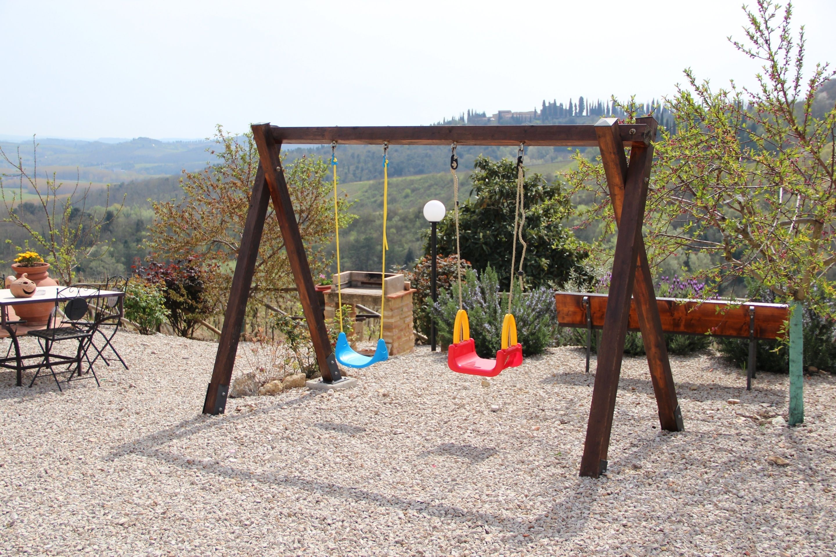 Un portique en bois installé dans un jardin, avec un sol recouvert de gravier, entouré d’arbres et offrant une vue sur des collines lointaines.