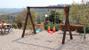 Un portique en bois installé dans un jardin, avec un sol recouvert de gravier, entouré d’arbres et offrant une vue sur des collines lointaines.