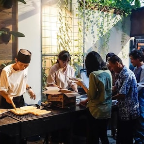 Un grupo de personas reunidas alrededor de un puesto de comida con una parrilla, una caja con el rótulo 'ENERGEN' y una pared con plantas trepadoras.