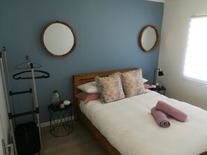 Cama bem arrumada com cabeceira de madeira, dois espelhos redondos, mesa de cabeceira com abajur e janela com persianas.