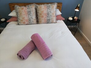 Cama bem arrumada com duas toalhas enroladas, cabeceira de madeira e mesa de cabeceira com abajur e objetos decorativos.
