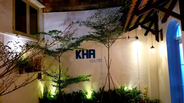 A cafe named "KHA" with a white wall, potted plants, and a small table. - KHA Hostel & Cafe (Ho Chi Minh City)
