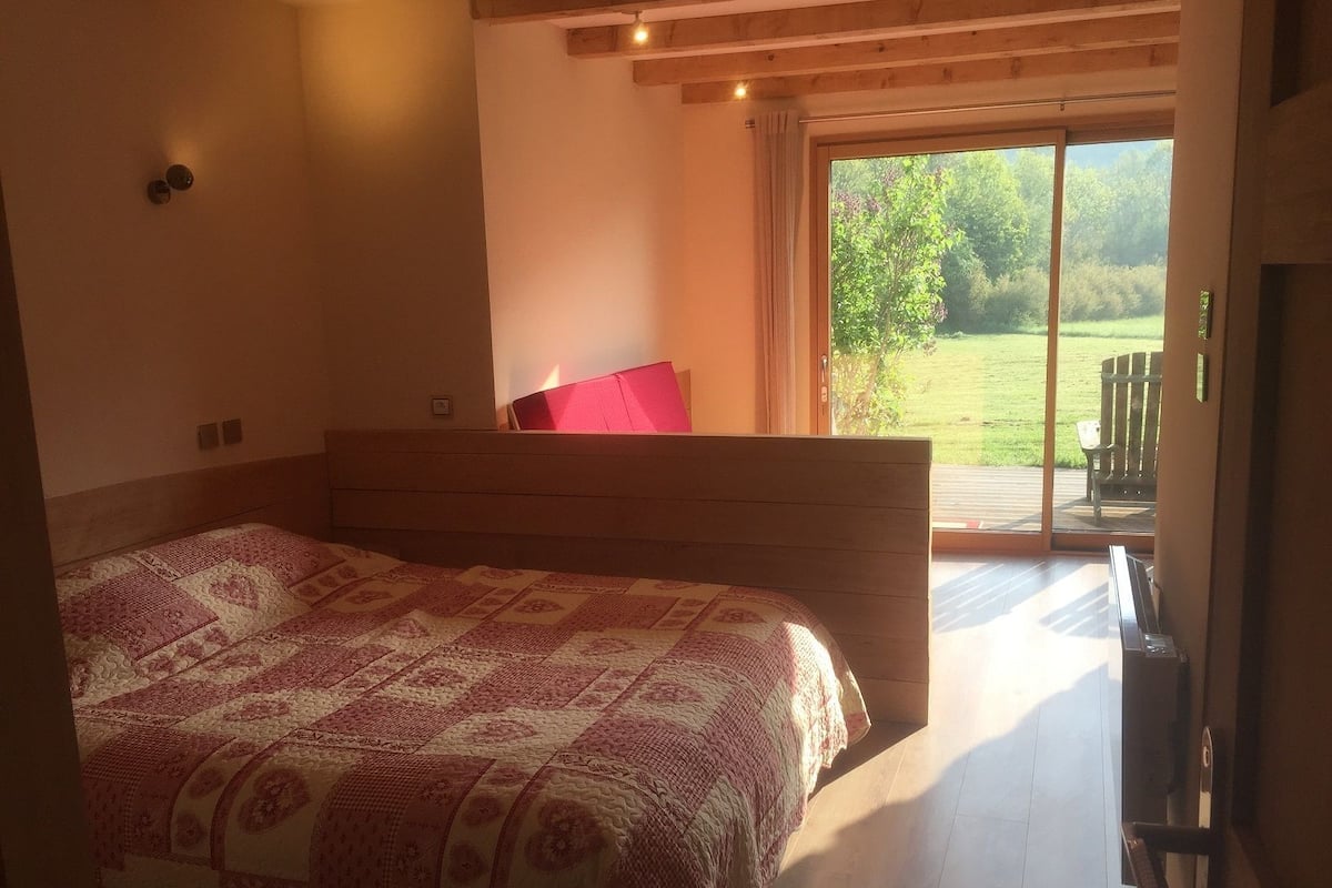 A bedroom with a large bed, a wooden headboard, a red pillow, and a sliding glass door leading to a balcony with a view of greenery.
