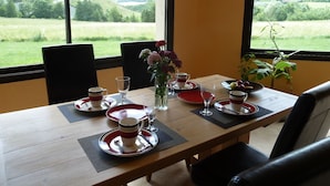 A dining table set with plates, cups, and a vase of flowers, with a view of a green landscape through large windows. - Alcove du Velay (Polignac)