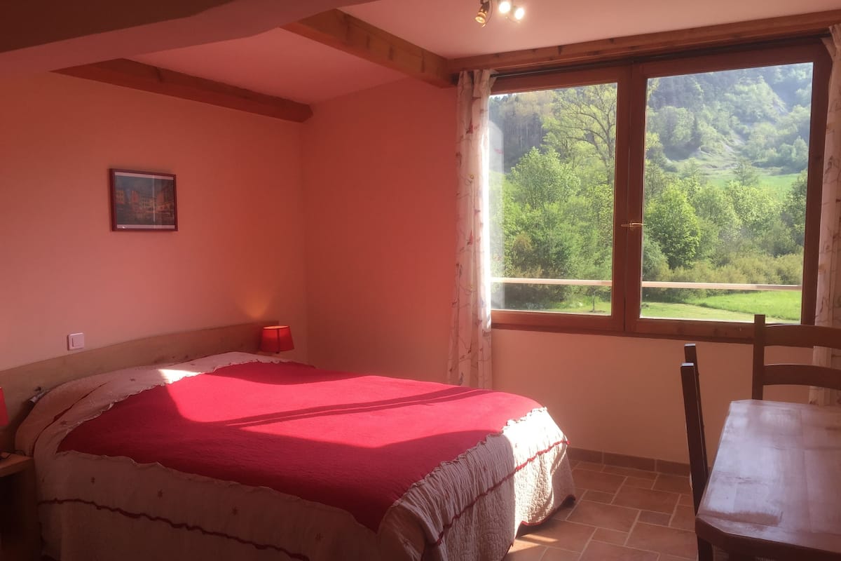 A bedroom with a bed, a table, a window with a view of greenery, and a painting on the wall.