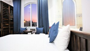 Habitación de hotel con una cama grande, dos ventanas con cortinas, una mesita con un florero y vistas a la ciudad al atardecer.