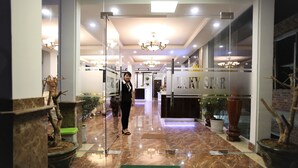 Un vestíbulo de hotel con un felpudo que dice 'BIENVENIDOS', plantas en macetas, un candelabro y un mostrador de recepción.