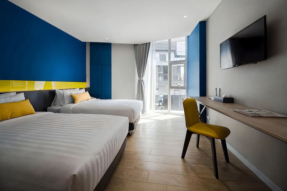 A hotel room with two beds, a desk, a chair, a TV, and a large window.