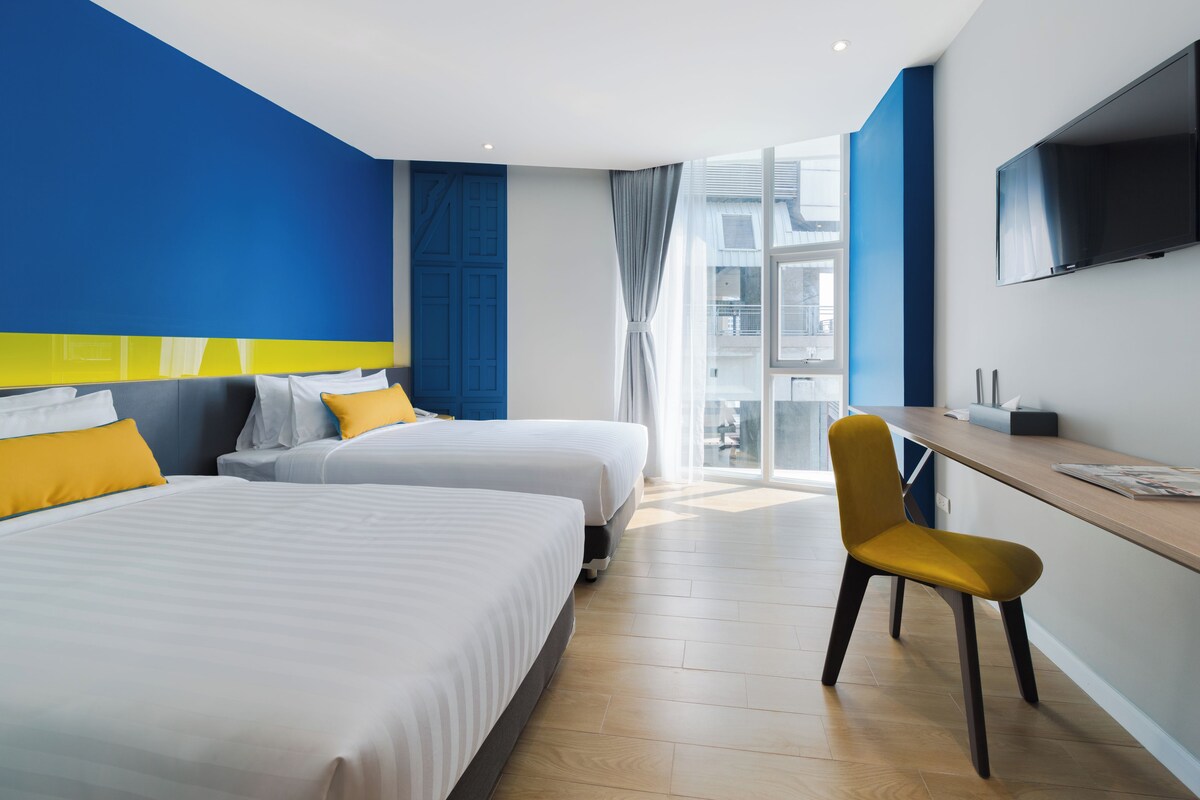 A hotel room with two beds, a desk, a chair, a TV, and a large window.