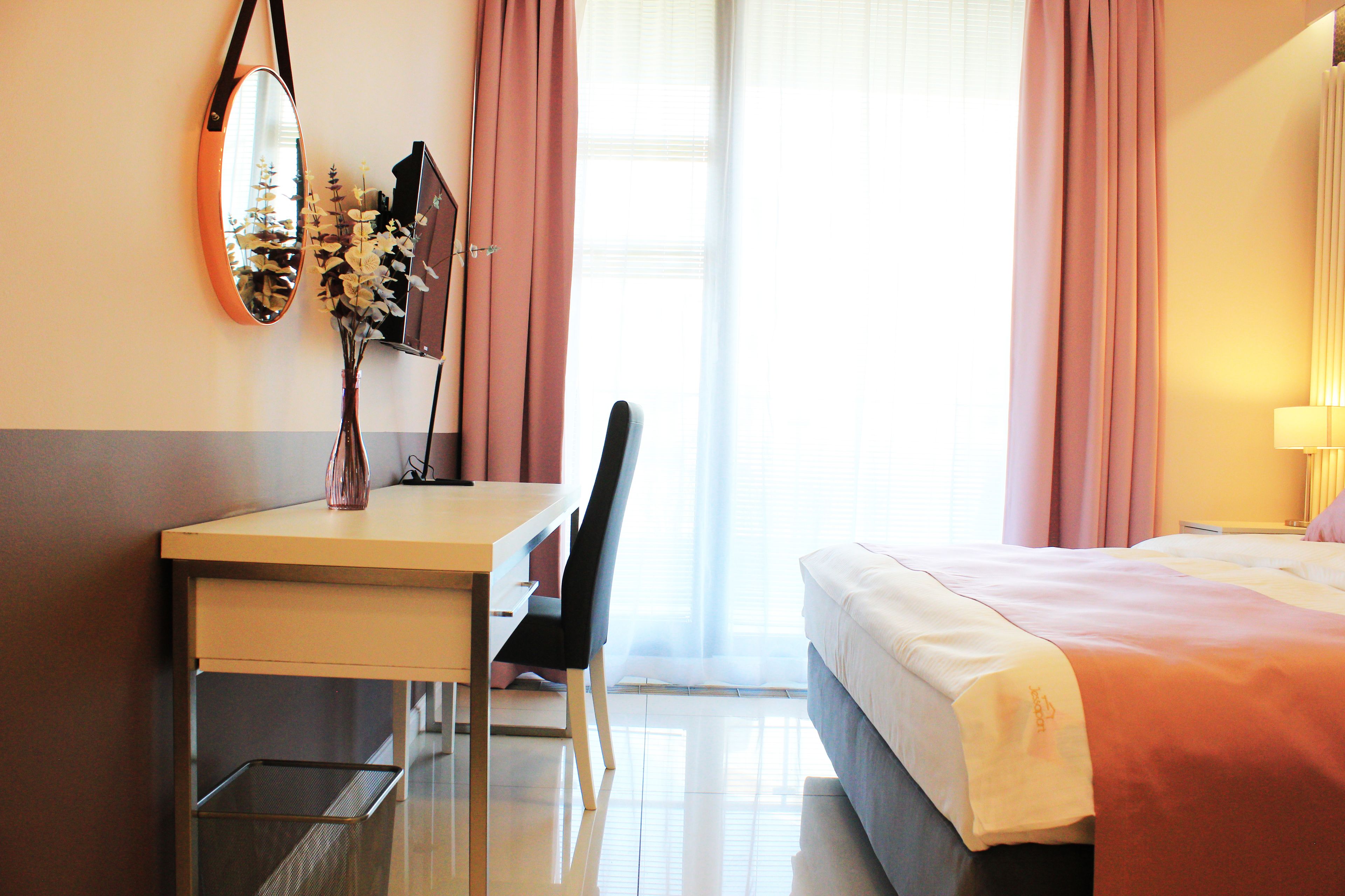 A hotel room with a bed, a desk with a chair, a mirror, and a vase with flowers.