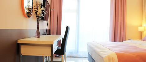 A hotel room with a bed, a desk with a chair, a mirror, and a vase with flowers.
