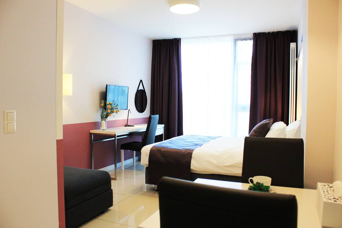 A hotel room with a bed, a desk, a chair, a TV, and a window with curtains.