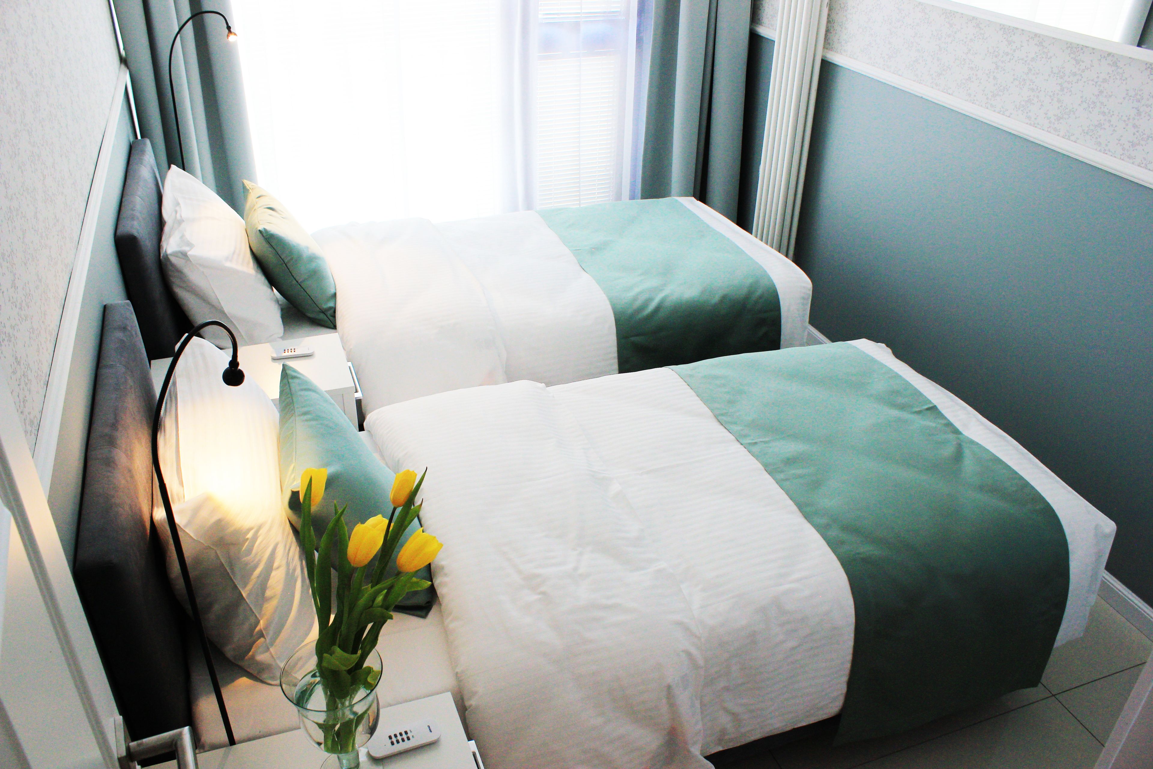 Une chambre d’hôtel avec deux lits, chacun recouvert d’une couvre-lit verte et blanche, une table de chevet avec une lampe et un vase rempli de fleurs jaunes.