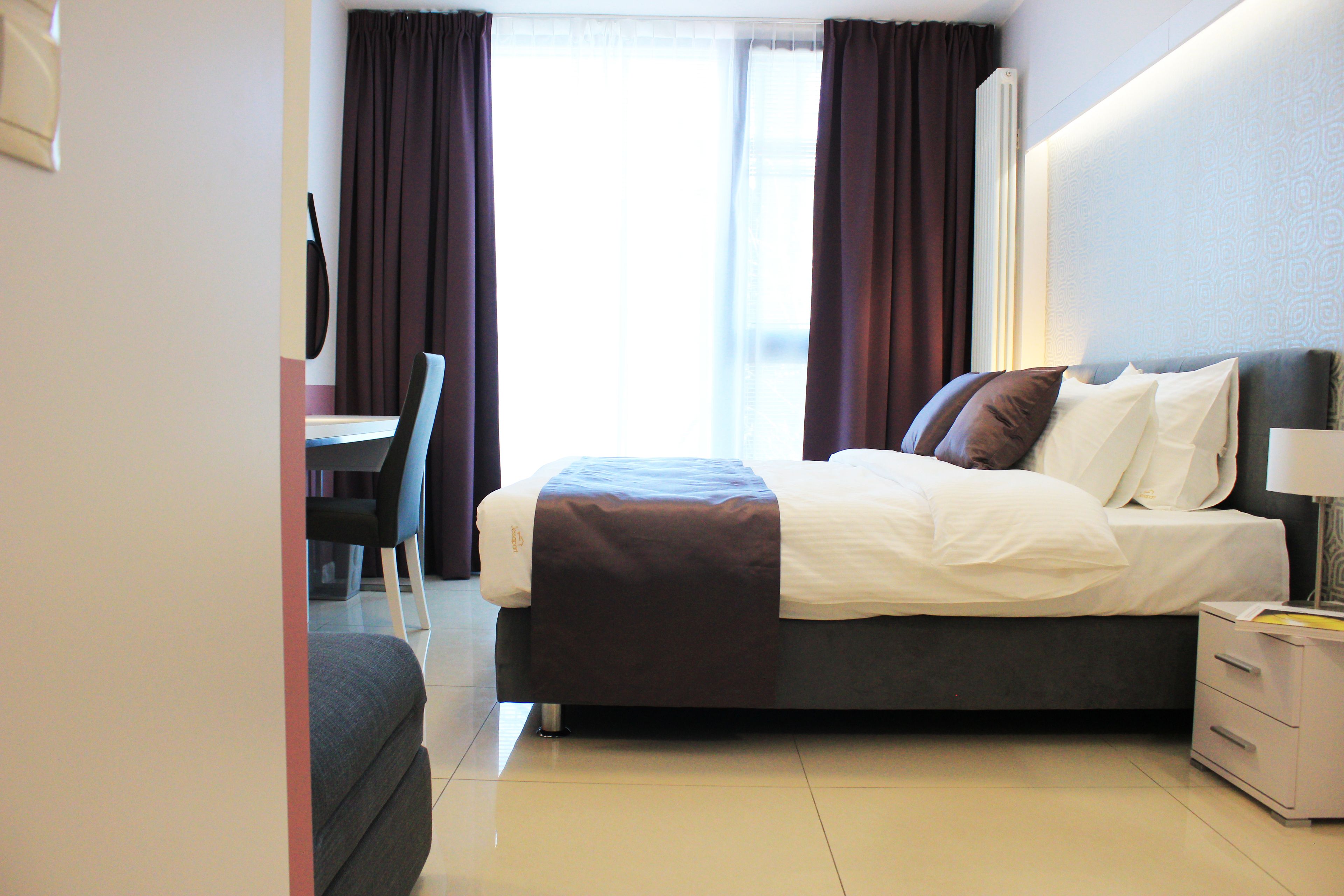 A hotel room with a bed, bedside table, chair, and a view of the outside through the window.