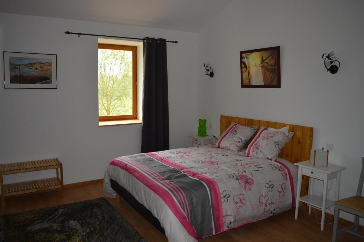 Un dormitorio con cama de madera, una mesita de noche, un banco, una ventana con cortinas y cuadros en la pared.