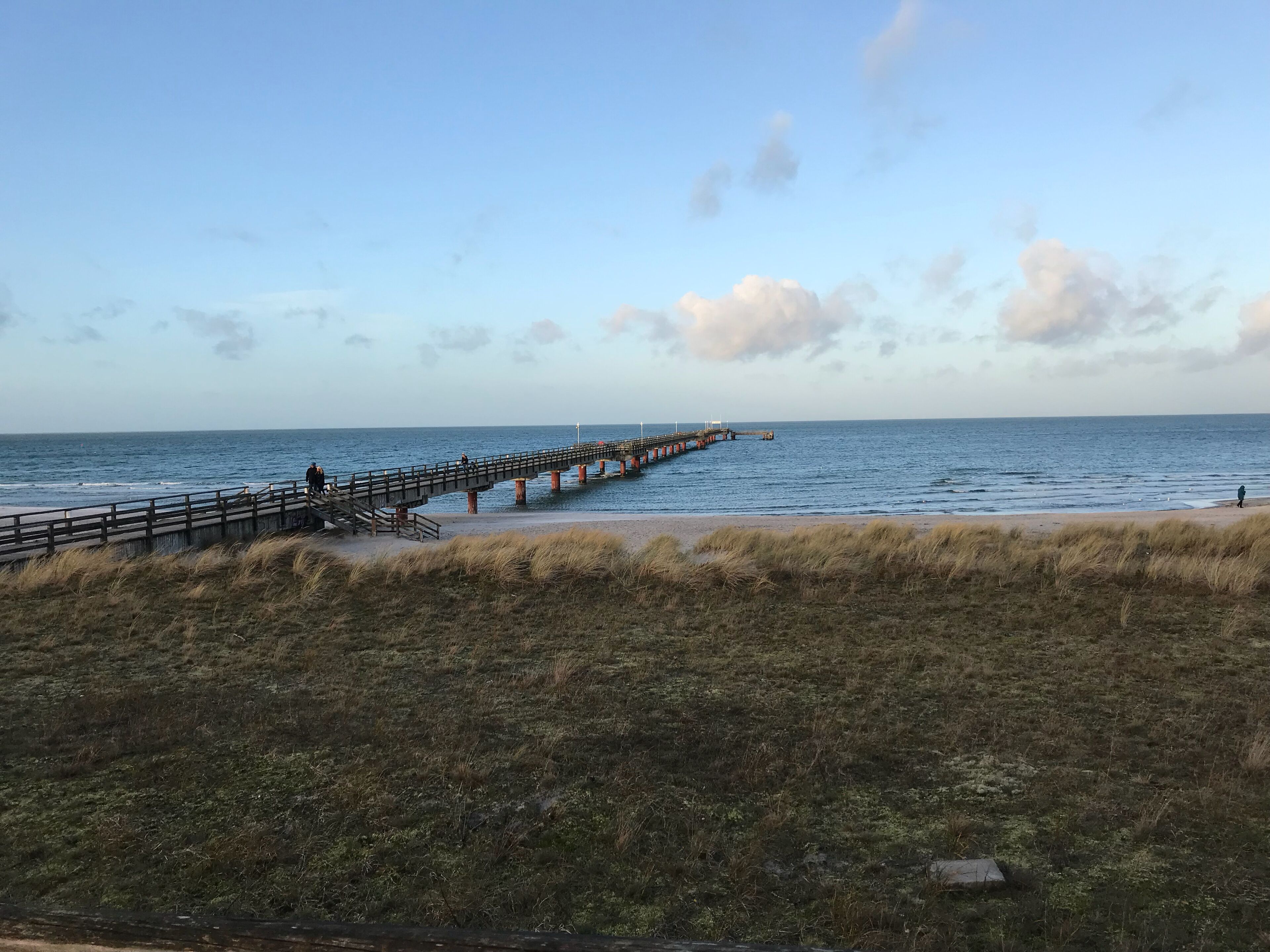 Un paysage côtier avec une jetée en bois s’avançant dans la mer, des dunes herbeuses au premier plan et un ciel dégagé.