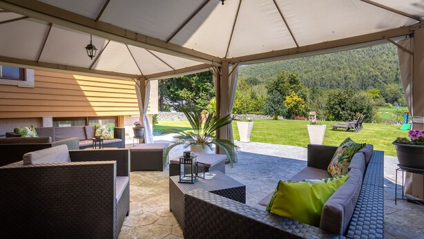 Un patio techado al aire libre con mobiliario de mimbre, plantas en macetas y vistas a un paisaje verde y exuberante.