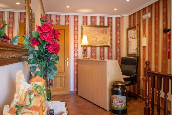 Ett väntrum med ett skrivbord i trä, en svart kontorsstol, en stor vas med röda blommor och en inramad tavla på väggen.