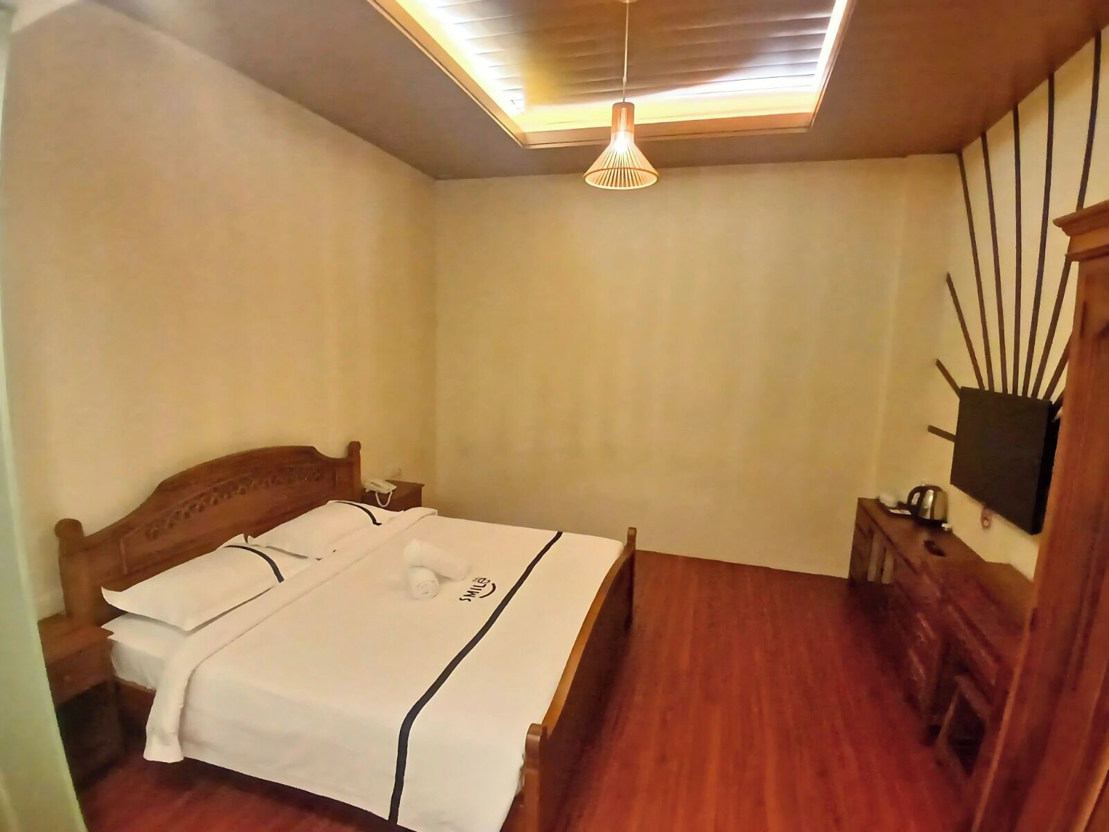 A bedroom with a wooden bed, a bedside table, a wall-mounted TV, and a ceiling light fixture.