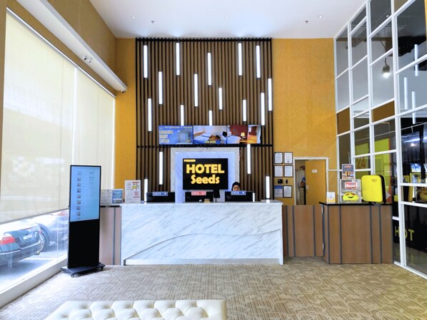 Un'area reception di un hotel con un bancone in marmo, una postazione self-service digitale e un grande schermo che mostra il nome dell'hotel.