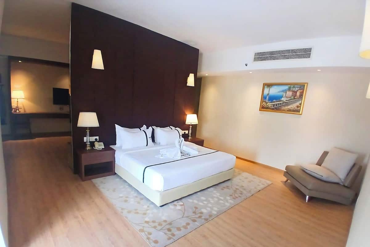 Ein Hotelzimmer mit einem großen Bett, Nachttischlampen, einem kleinen Schreibtisch, einem Sessel und einem Bild an der Wand.