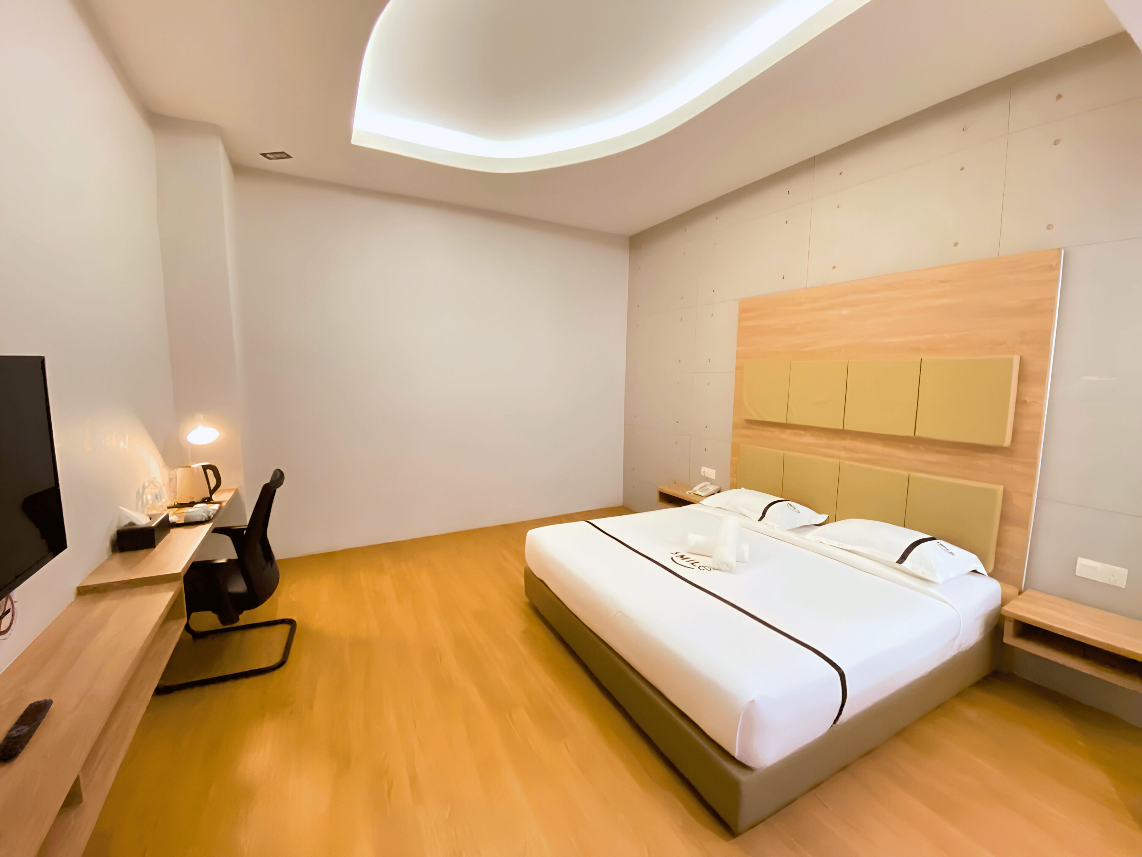 A modern hotel room with a large bed, a desk with a chair, a flat-screen TV, and a bedside table.